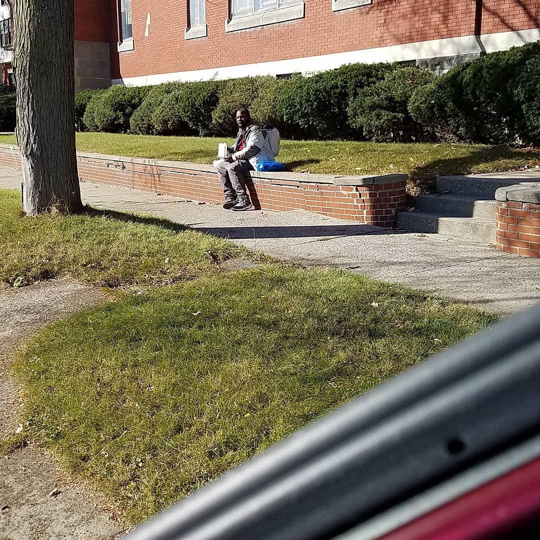 Person sitting on a brick planter beside a hedge in front of a brick building.