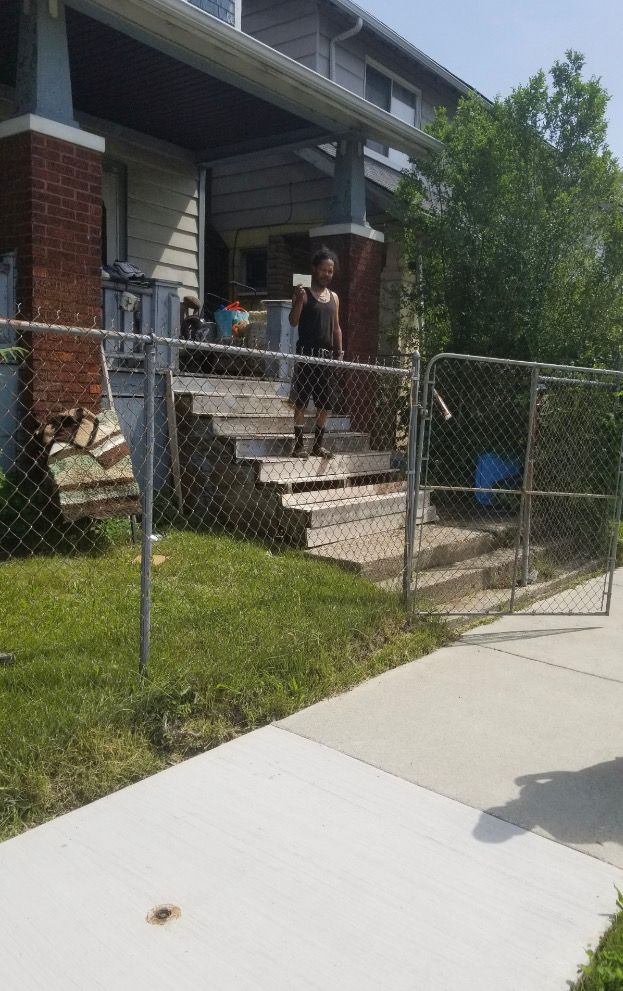 Two people standing on the front steps of a brick house behind a chain-link fence, with a sidewalk in front