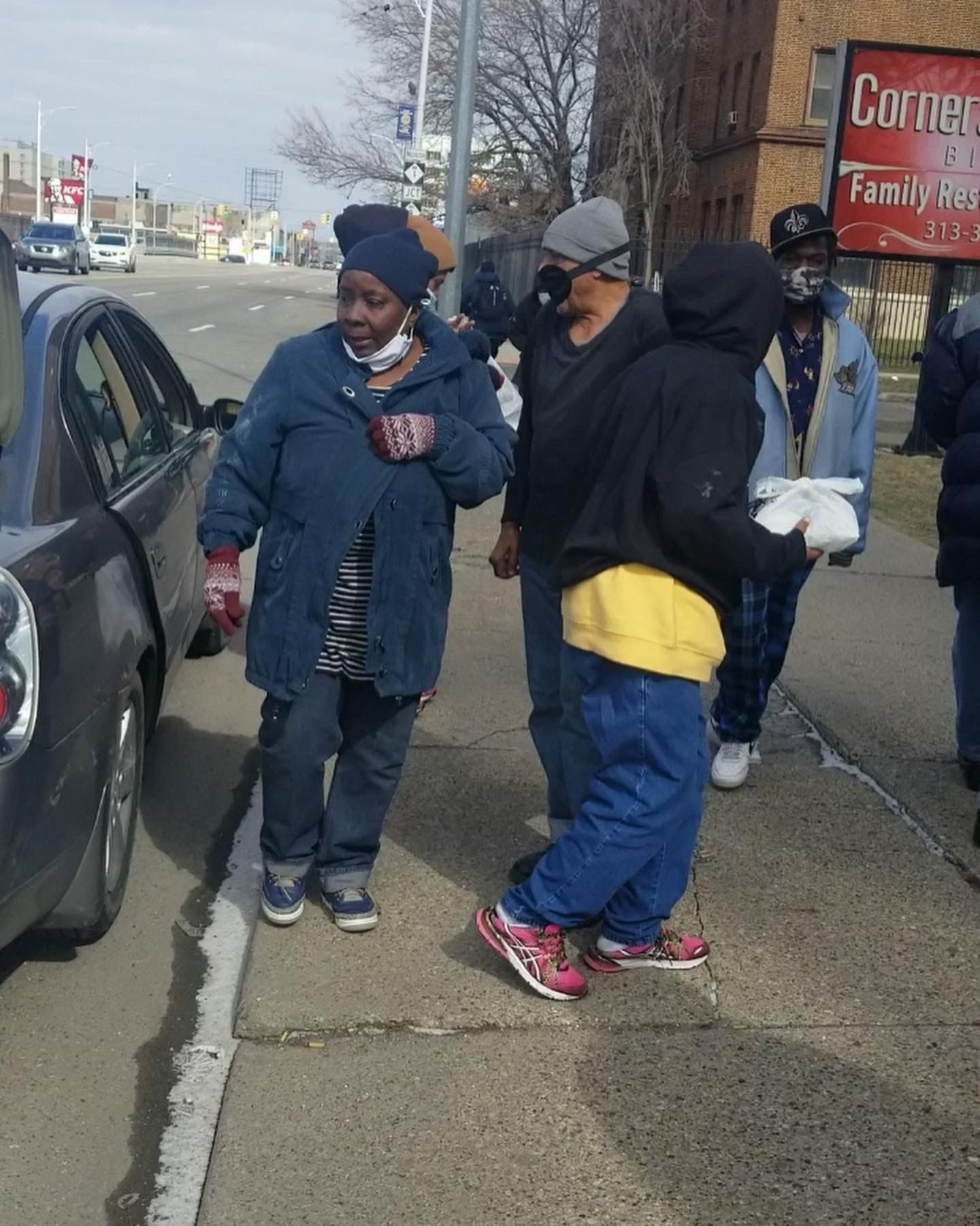 Three people in winter coats stand on a sidewalk beside a parked car.