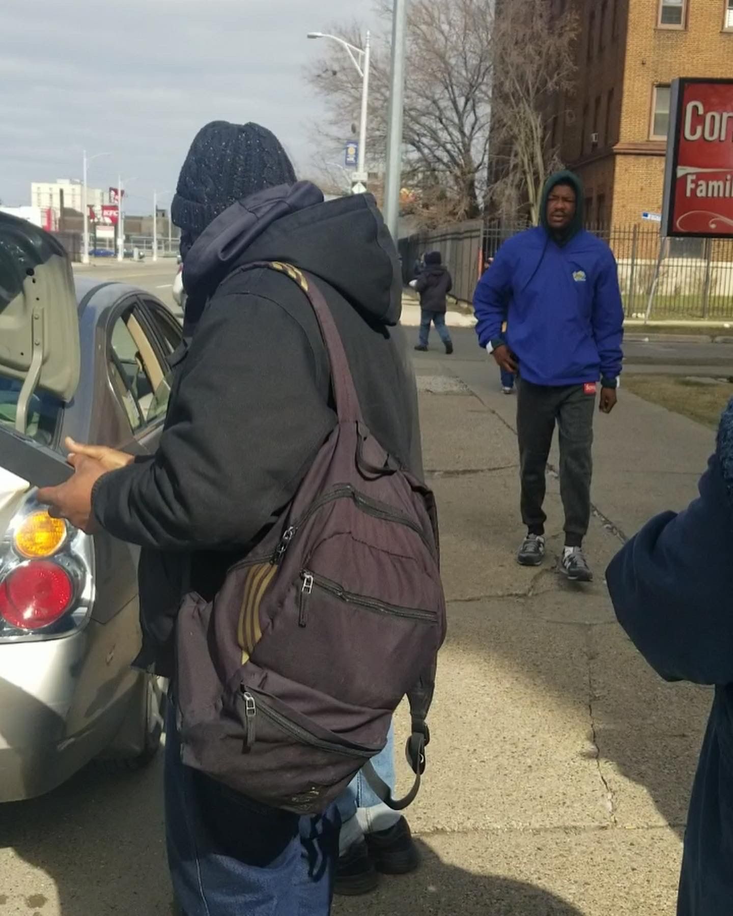 People standing beside a parked car on a city sidewalk, one with a backpack, another in a blue jacket.