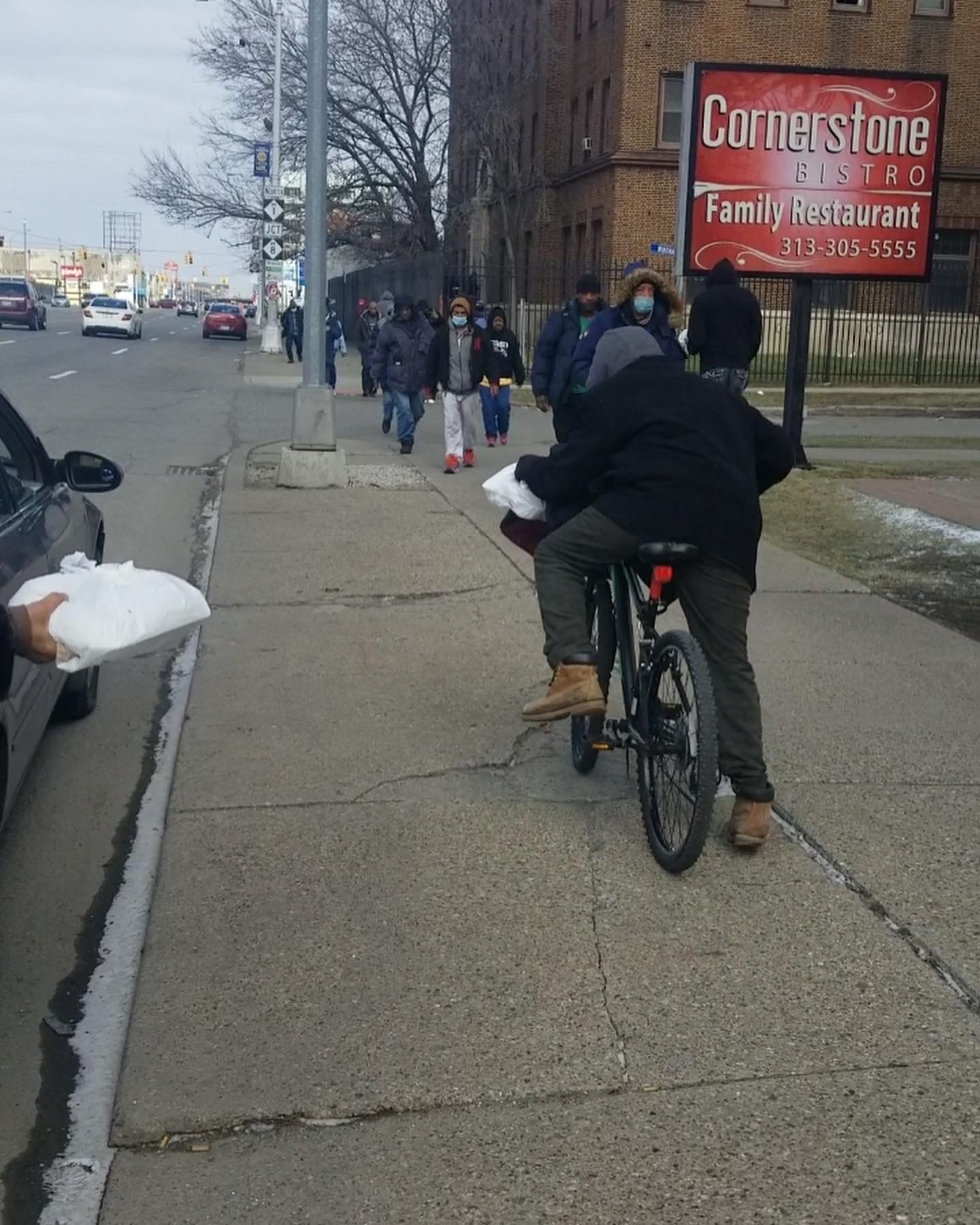 Person on bicycle on a city sidewalk beside a red Cornerstone Family Restaurant sign, with pedestrians ahead.