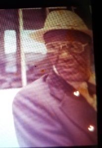 Man wearing a cap and glasses, seated indoors beside a window, in a purple shirt.