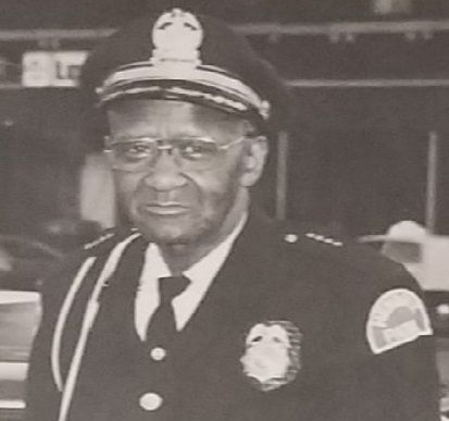Uniformed police chief in a peaked cap and glasses, standing indoors and facing the camera.