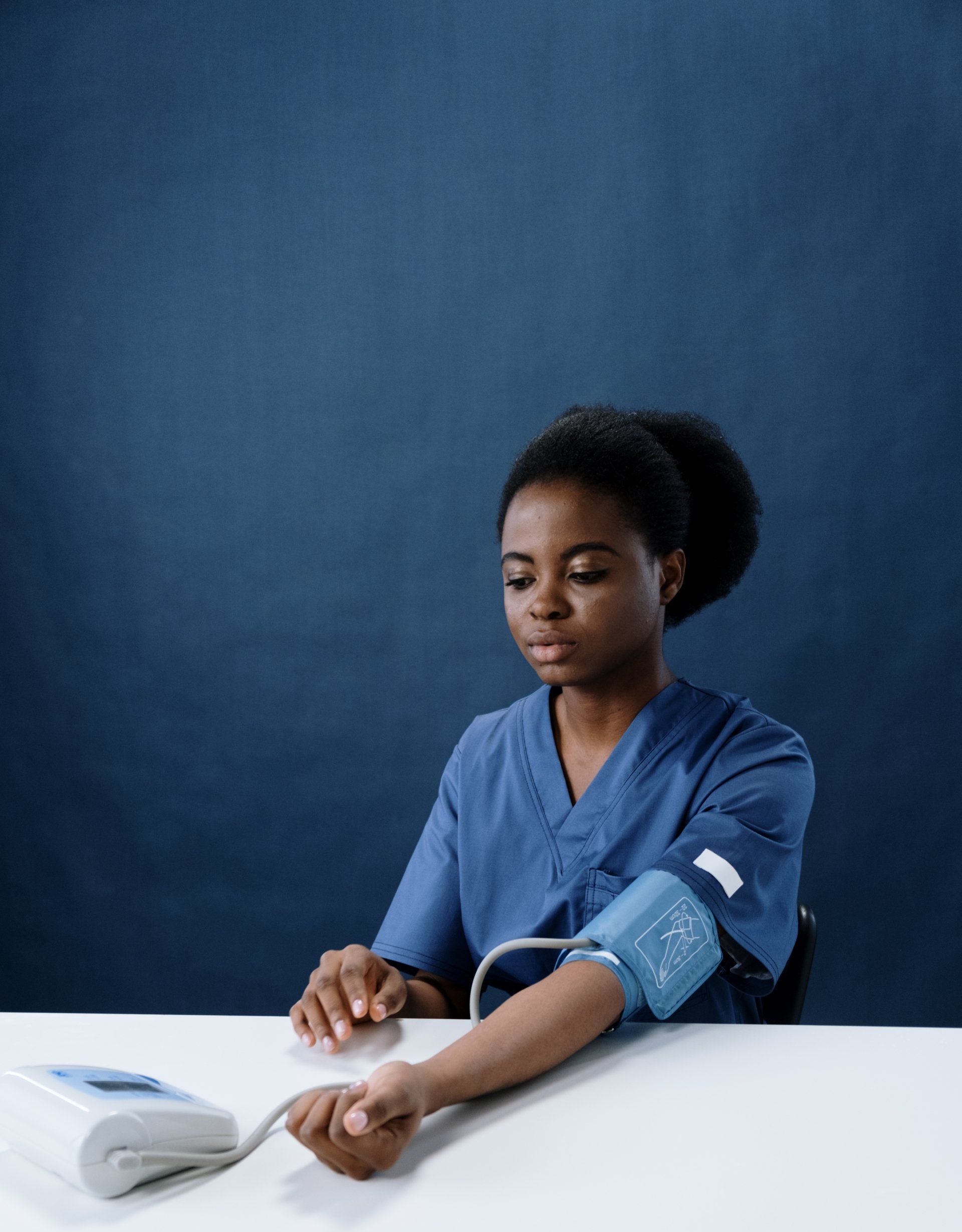 Person in blue scrubs checking blood pressure with a cuff on a white table against a blue backdrop