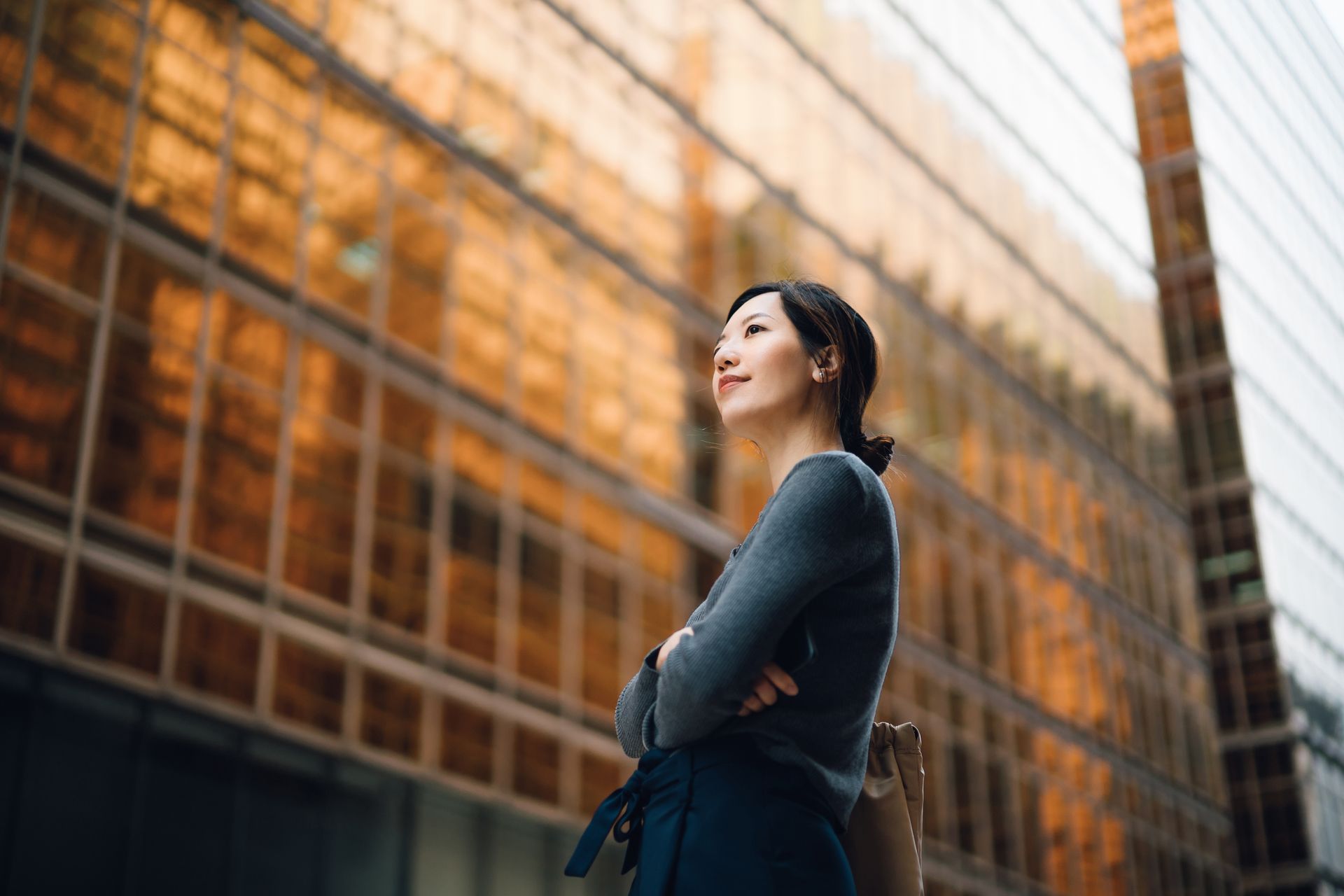Woman with arms crossed, looking up, stands in front of a gold-tinted glass building.