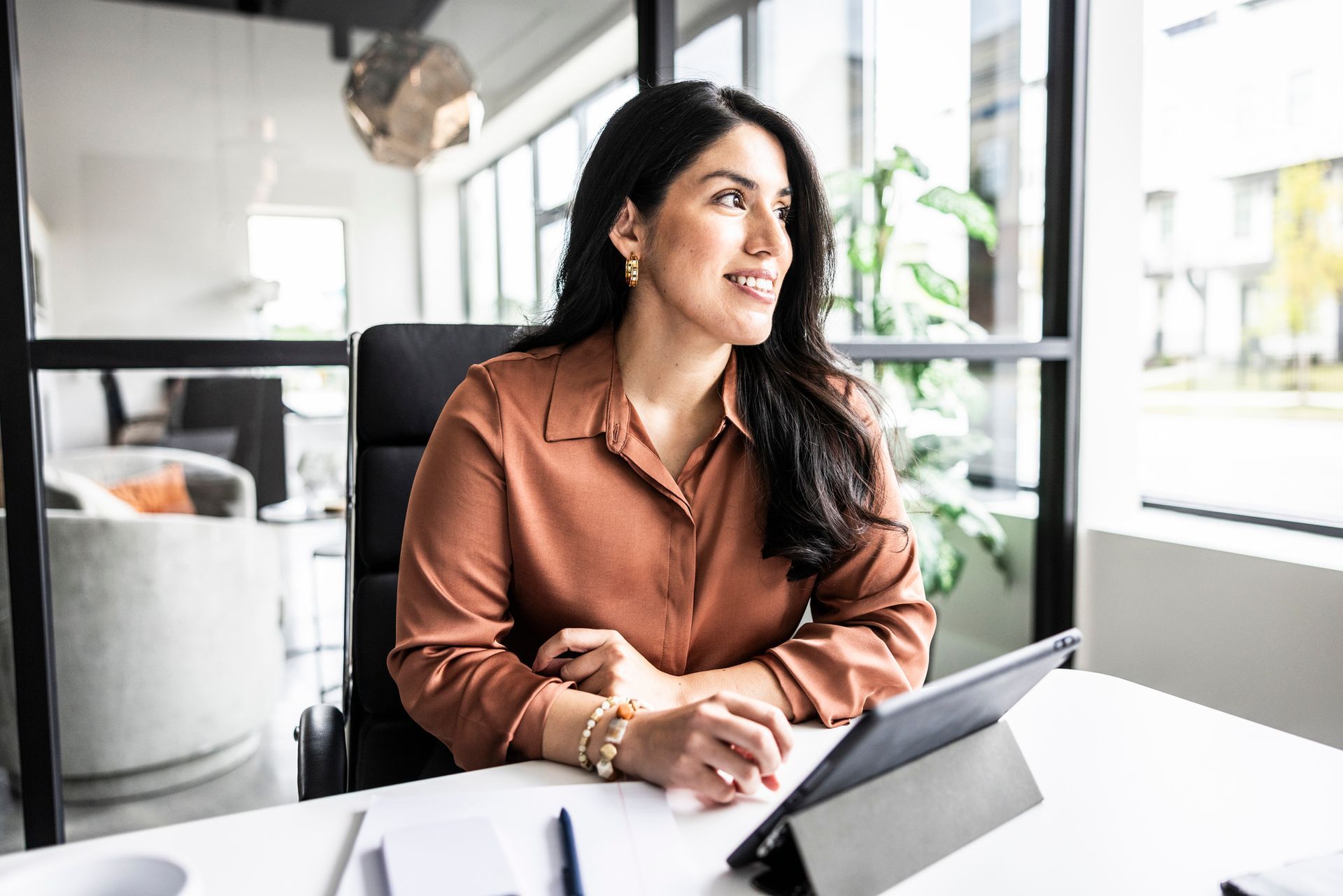 Woman in brown shirt, looking out window at desk with tablet, smiling. Office setting.