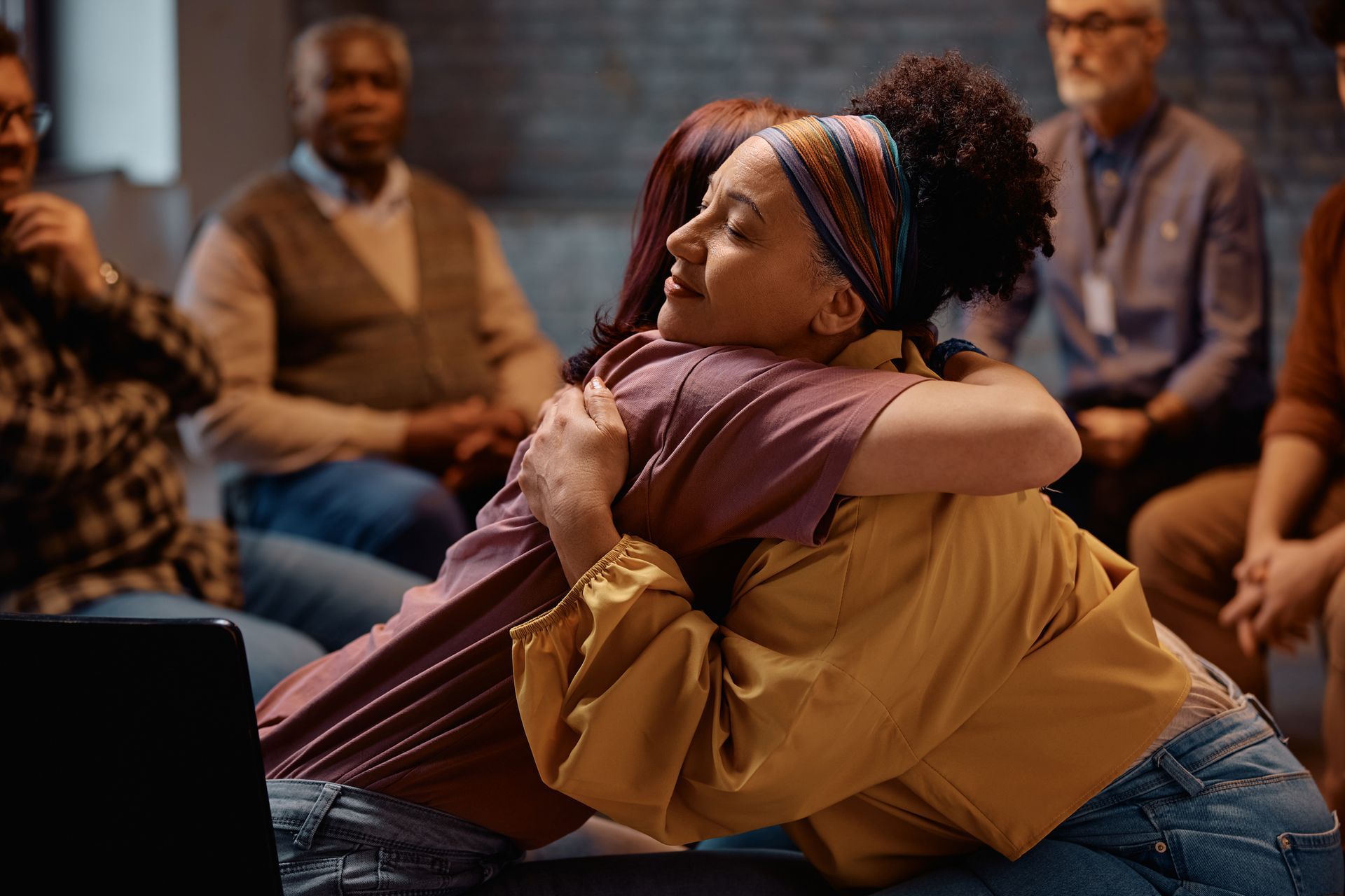 Two women embracing in support group; others seated in circle.