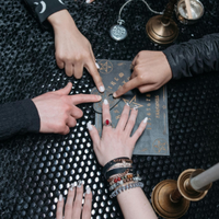 Hands pointing at a board game or ritual board on a table, surrounded by candles and jewelry.