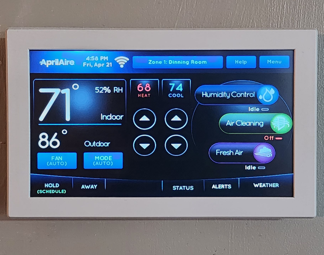 Wall-mounted AprilAire thermostat touchscreen showing indoor/outdoor temperatures and humidity controls