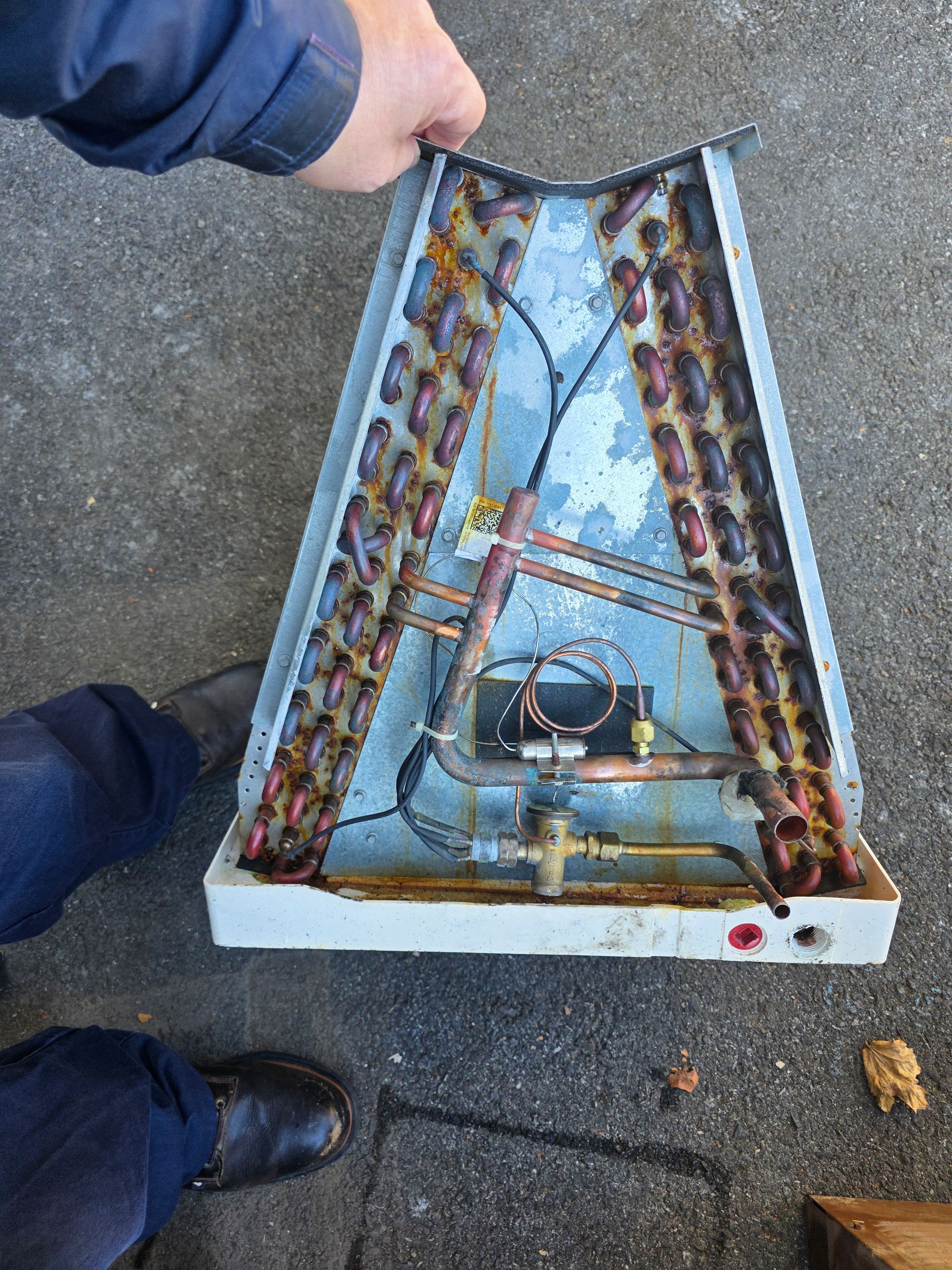 Open metal box with wiring and tubing, held above pavement by a person’s hand and feet visible nearby