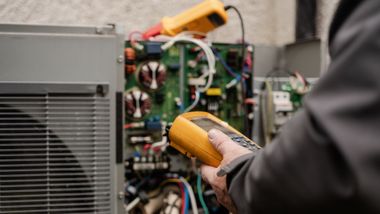 Technician using a yellow multimeter to test an open electrical panel with exposed wiring