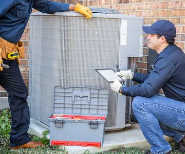 Two HVAC technicians inspecting an outdoor air conditioner beside a brick wall with a toolbox on the ground