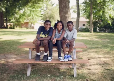 Three children sit on a picnic table outdoors, smiling, in a sunny park.