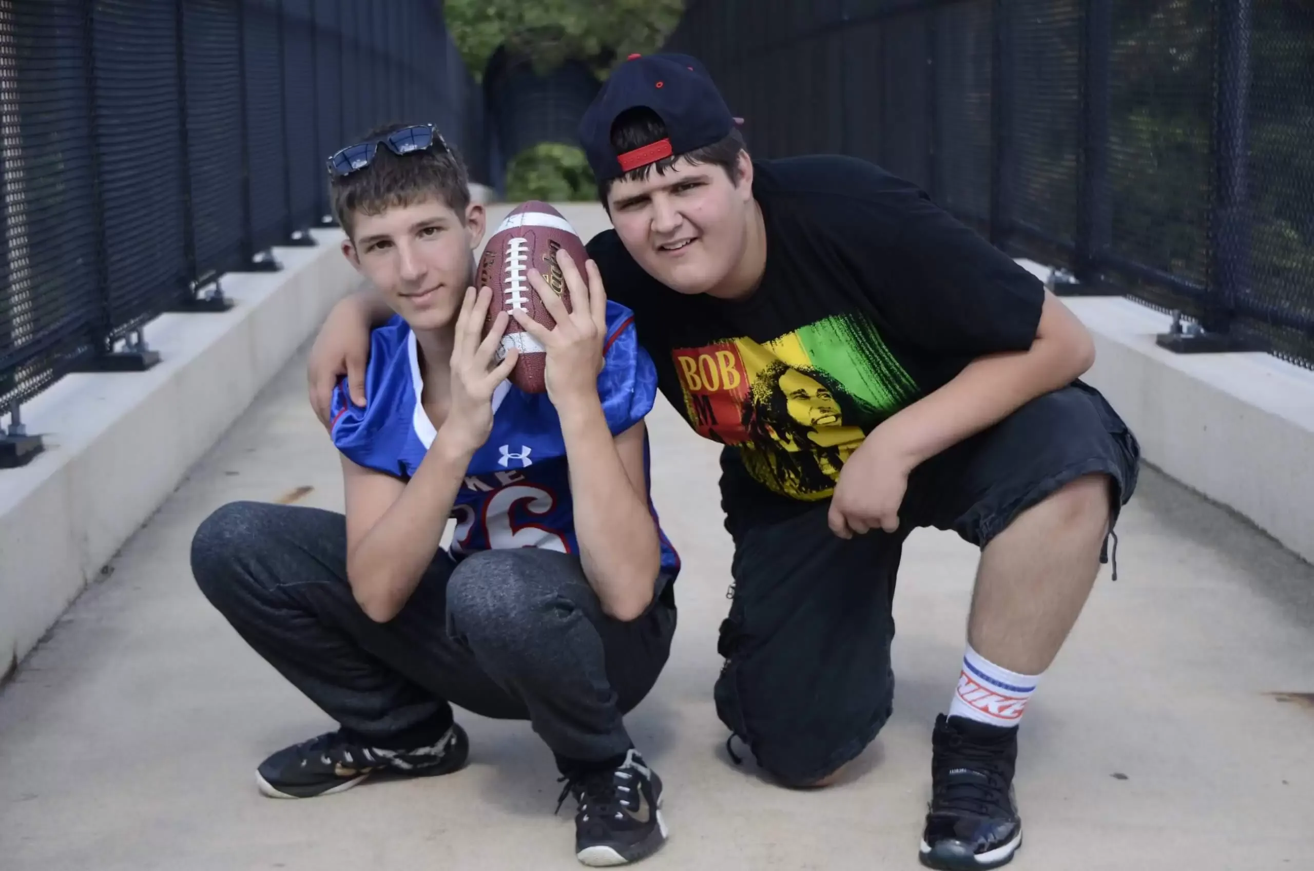 Two young men pose on a bridge with a football. One squats, holding the ball; the other has his arm around him.