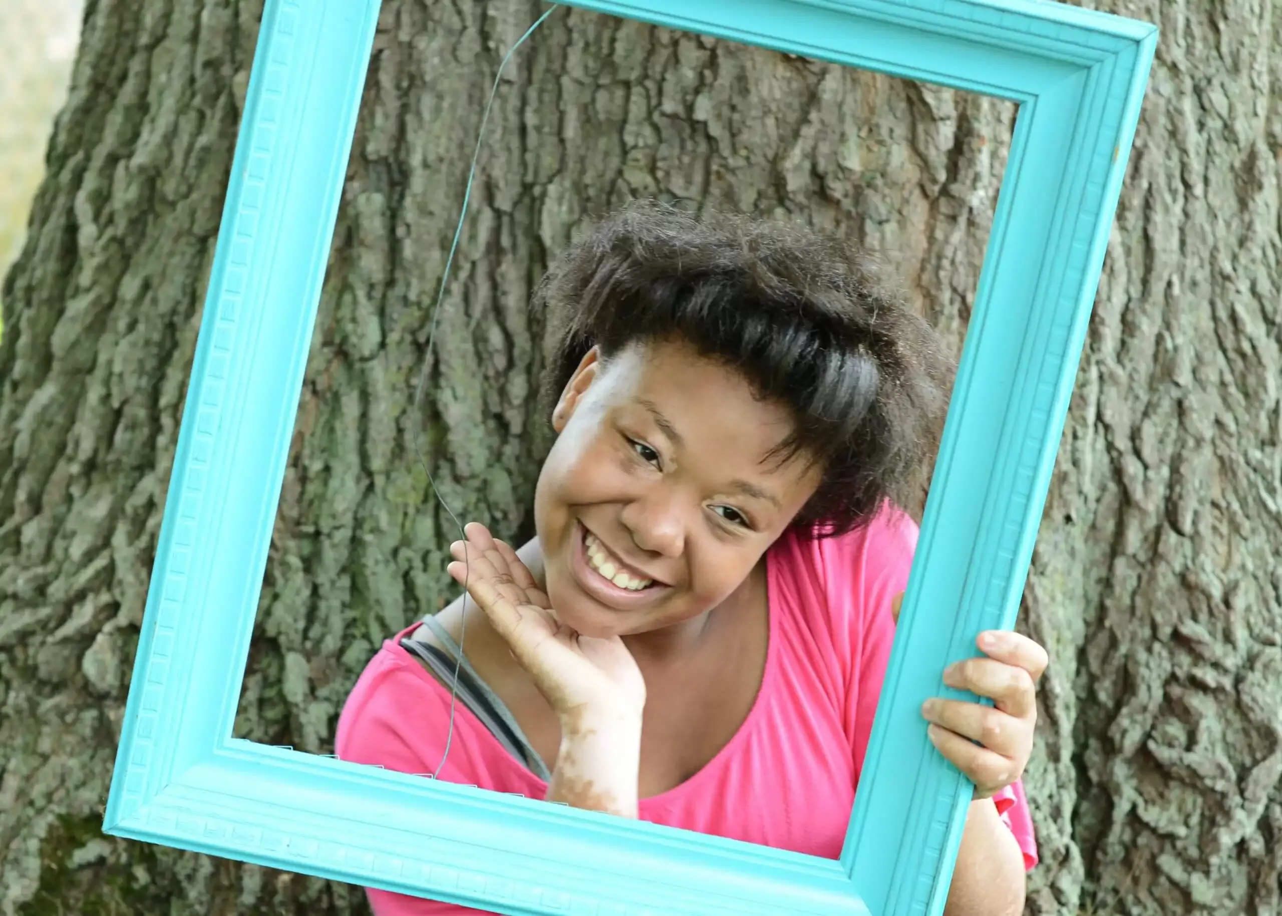 Smiling person framed by a teal square, set against a tree; pink shirt, dark hair.