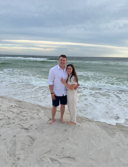 Couple standing on a sandy beach, man in shorts and white shirt, woman in tan jumpsuit, ocean in background.