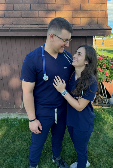 A man and woman in navy scrubs gaze at each other, smiling, with a shed and greenery in the background.