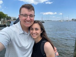 A smiling couple takes a selfie near water with boats. The man wears glasses, the woman has a backpack.