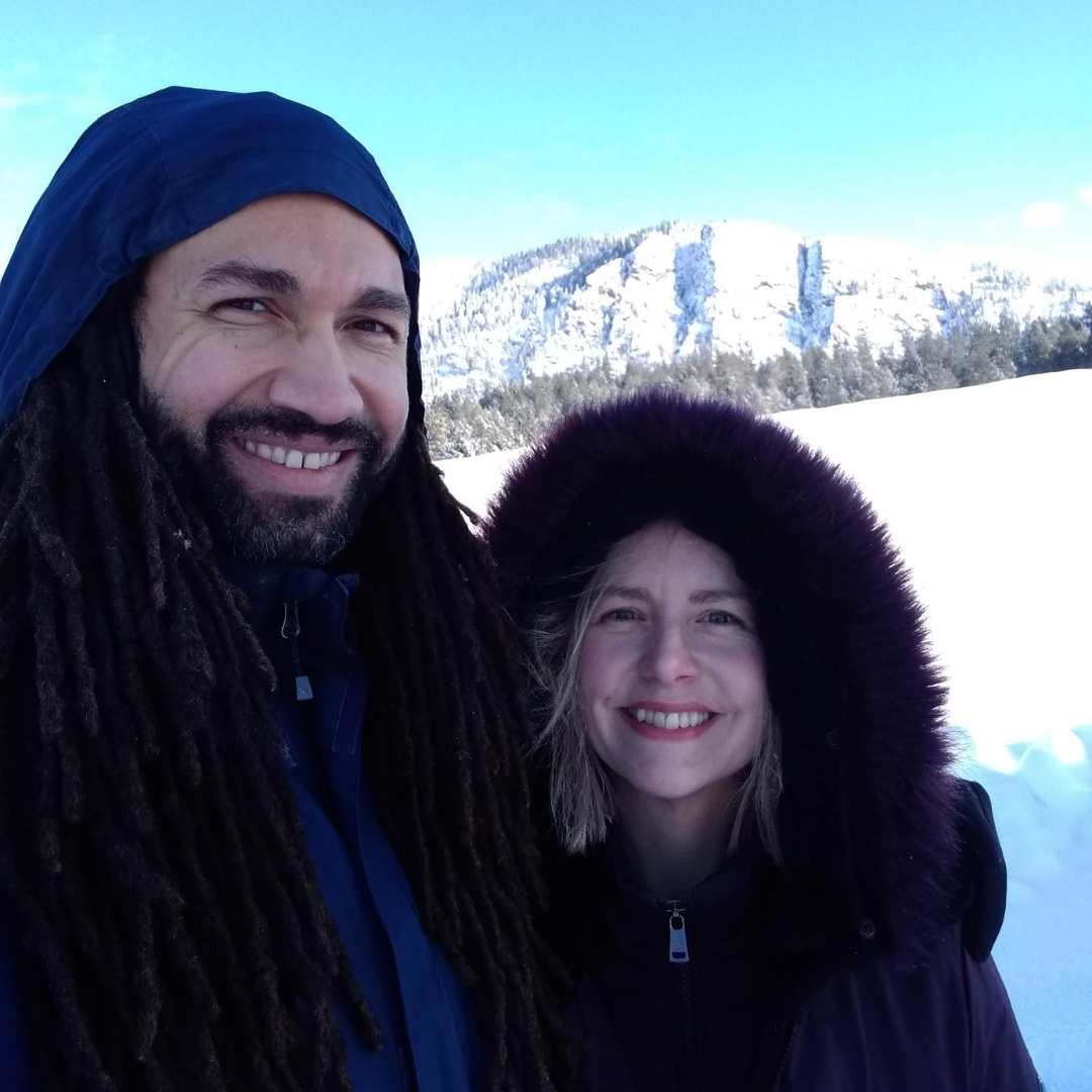 Couple smiling in snowy landscape, man with dreadlocks and blue jacket, woman in hooded coat.