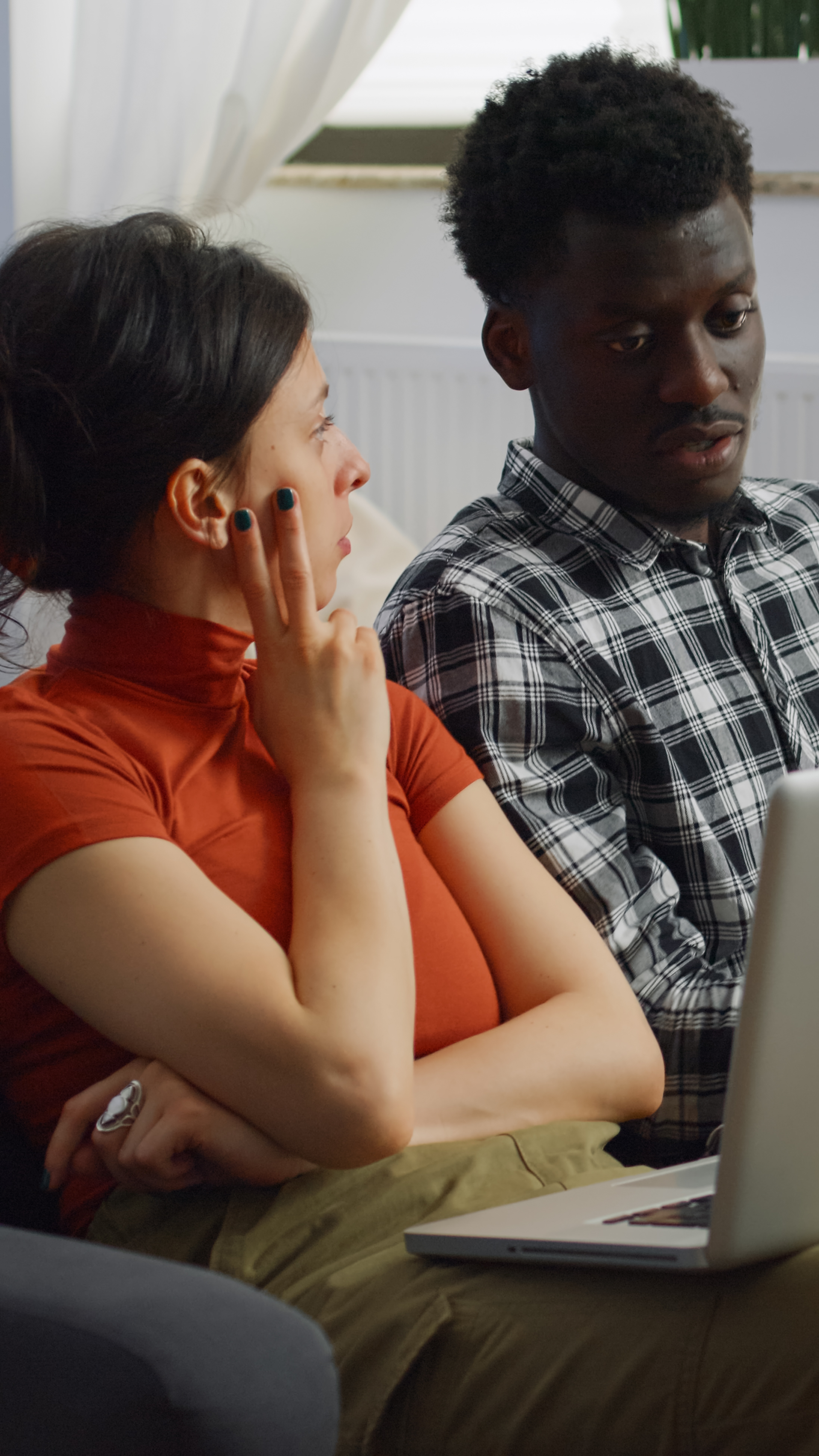 A diverse couple looking at a laptop, woman with hand to face, man with plaid shirt, indoors.