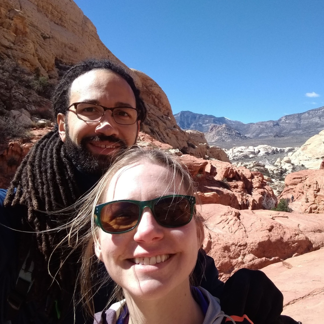 Couple smiling in front of red rock mountains under a blue sky.