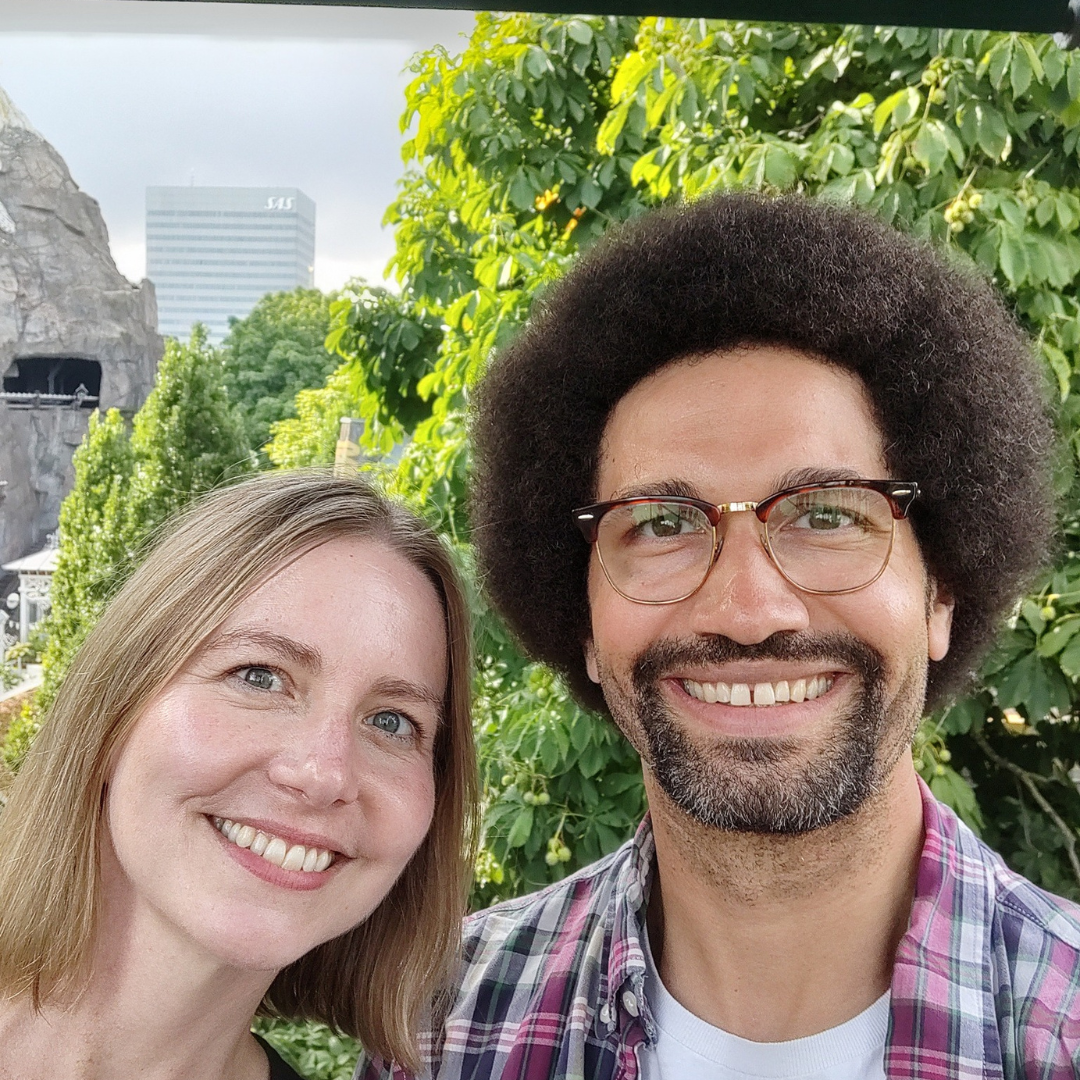 Couple smiling for a selfie outdoors; woman with blonde hair, man with afro and glasses; trees, building in background.