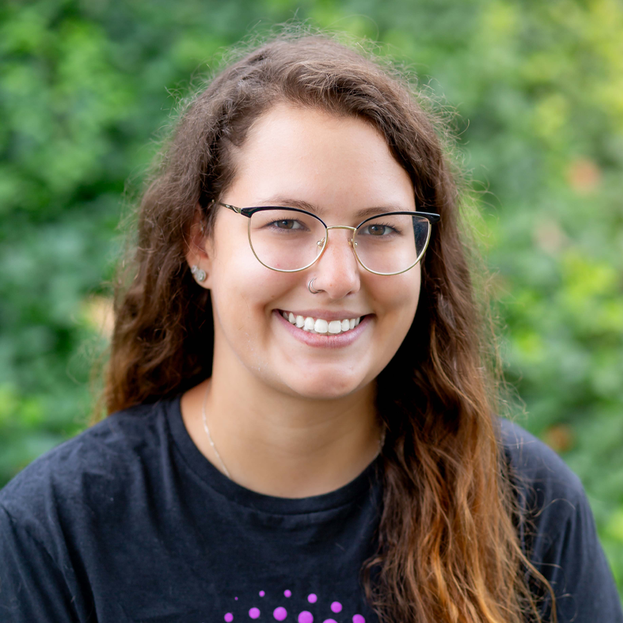 Woman with glasses smiles, wearing a dark shirt, in front of green foliage.