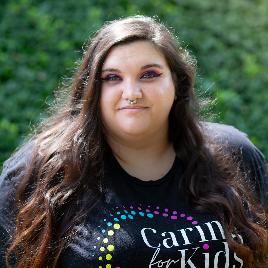 Woman with long brown hair and nose ring smiles, wearing a black t-shirt that says 