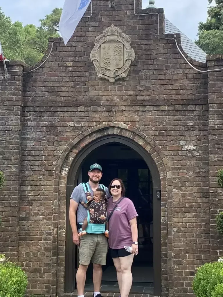 Family poses in front of a brick building entrance. Man holds a baby in a carrier; woman stands beside them.
