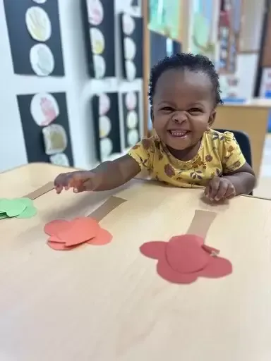 Smiling child at a table with colorful paper cutouts. Classroom setting, happy expression.