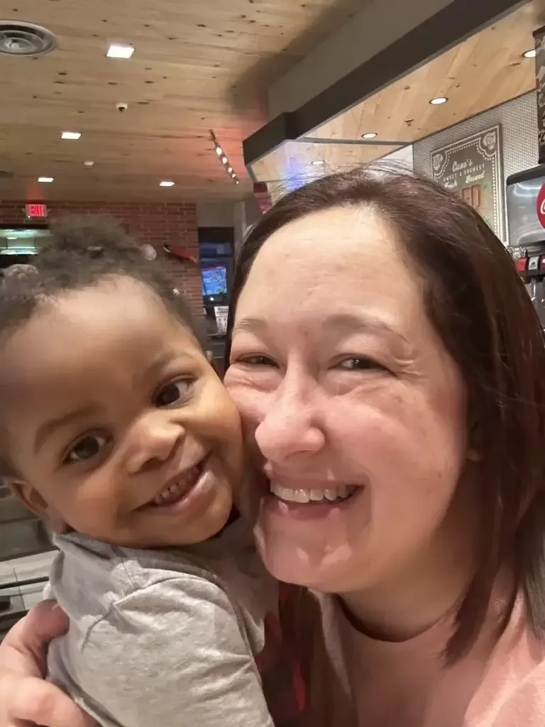 Woman smiling, hugging a smiling baby. Indoor setting, warm lighting.