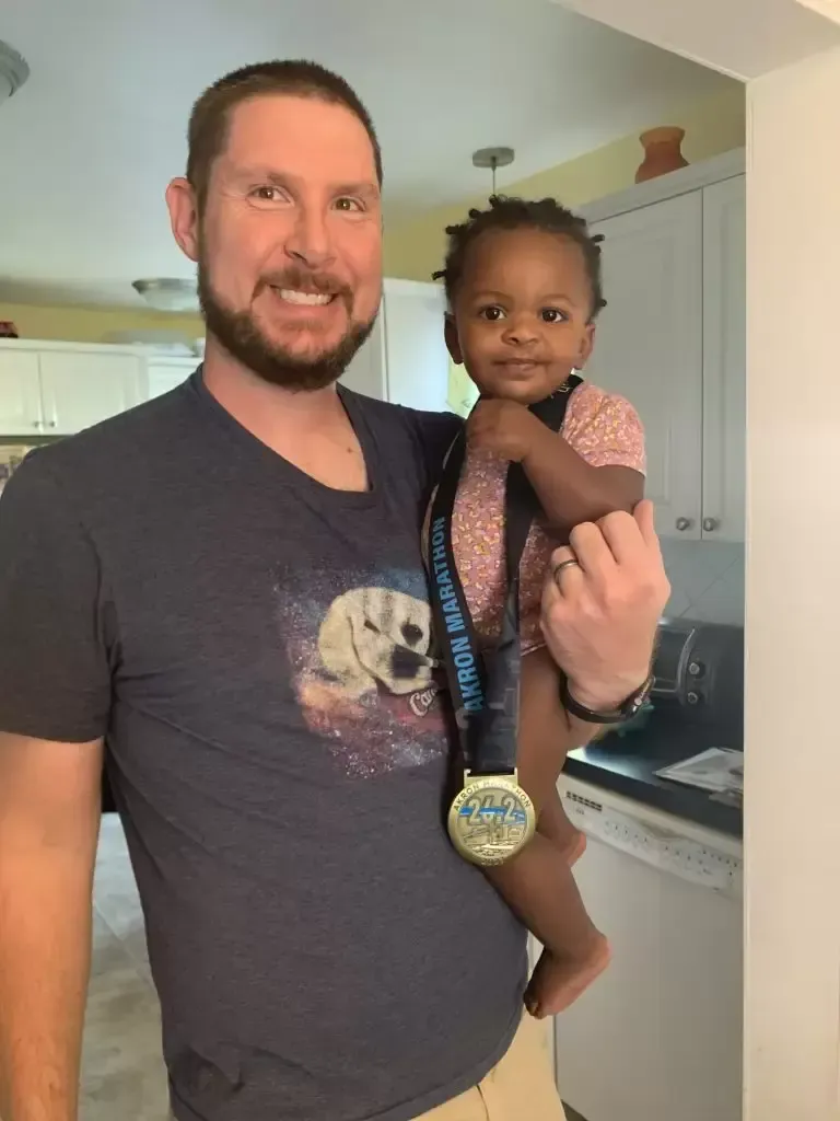 Man holding baby, both smiling; man wears shirt with dog graphic, baby has medal.