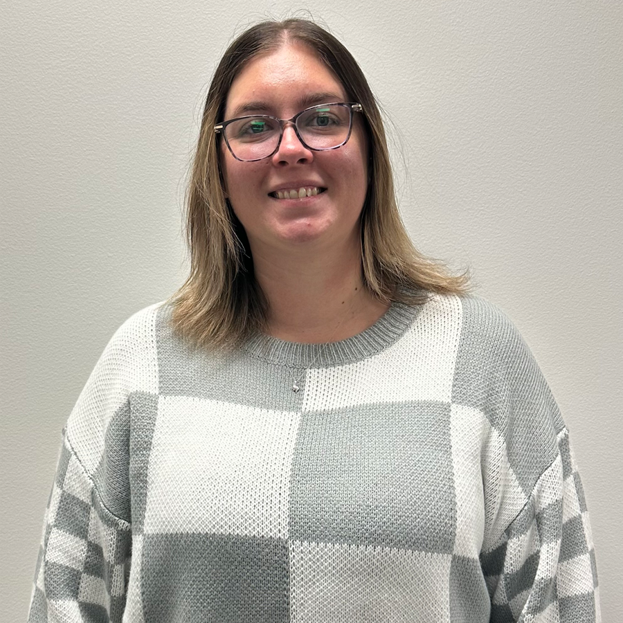 Woman with glasses, wearing a gray and white checkered sweater, smiling in front of a white wall.