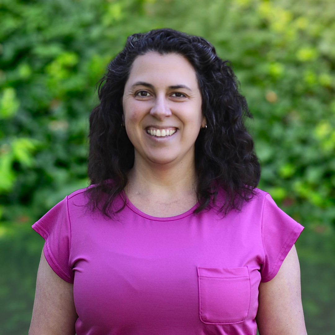 Woman with dark curly hair smiles broadly, wearing a pink long-sleeved shirt, standing against a grey backdrop.