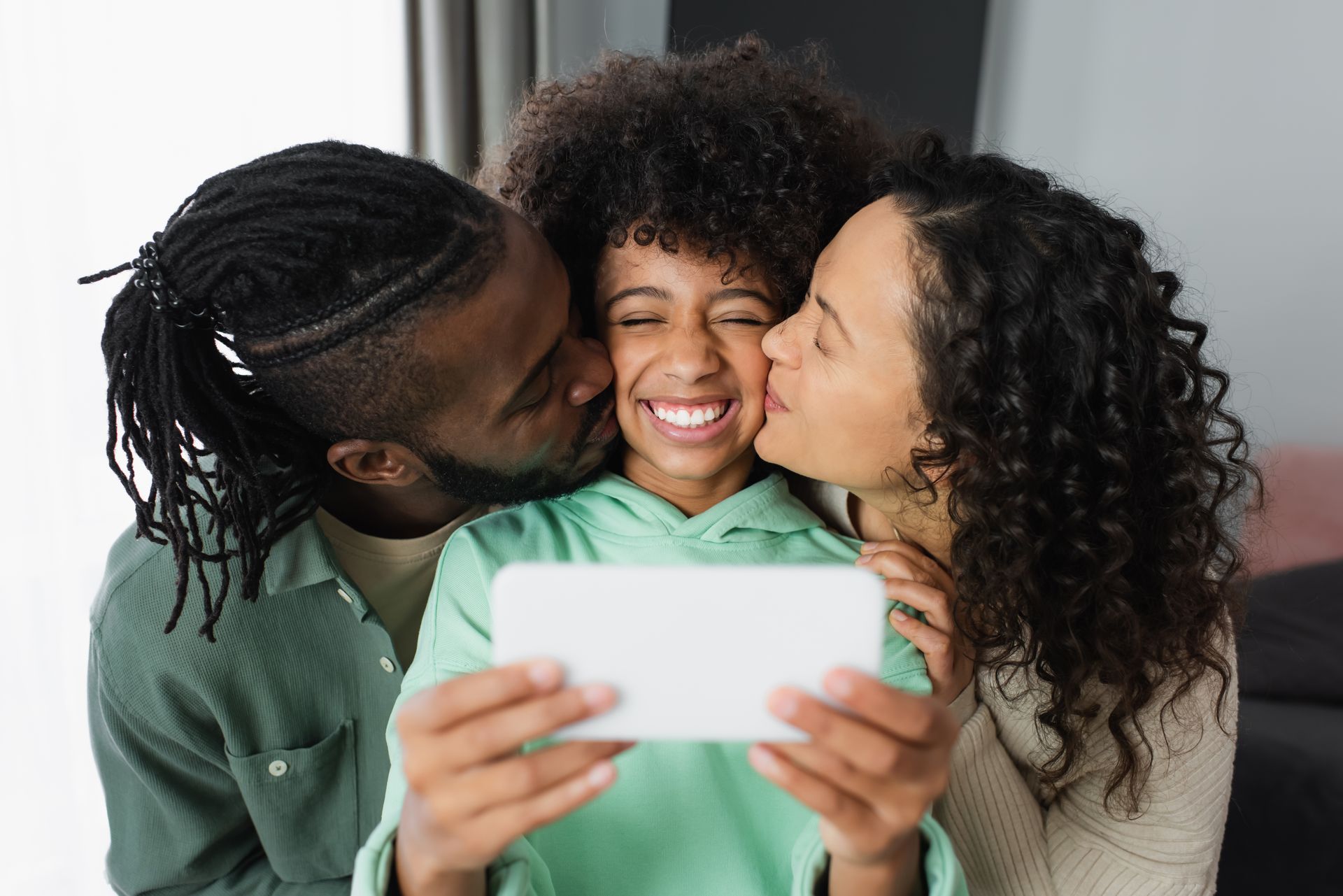Family takes a selfie, smiling. One parent kisses the child's cheek, the other parent kisses the other cheek.