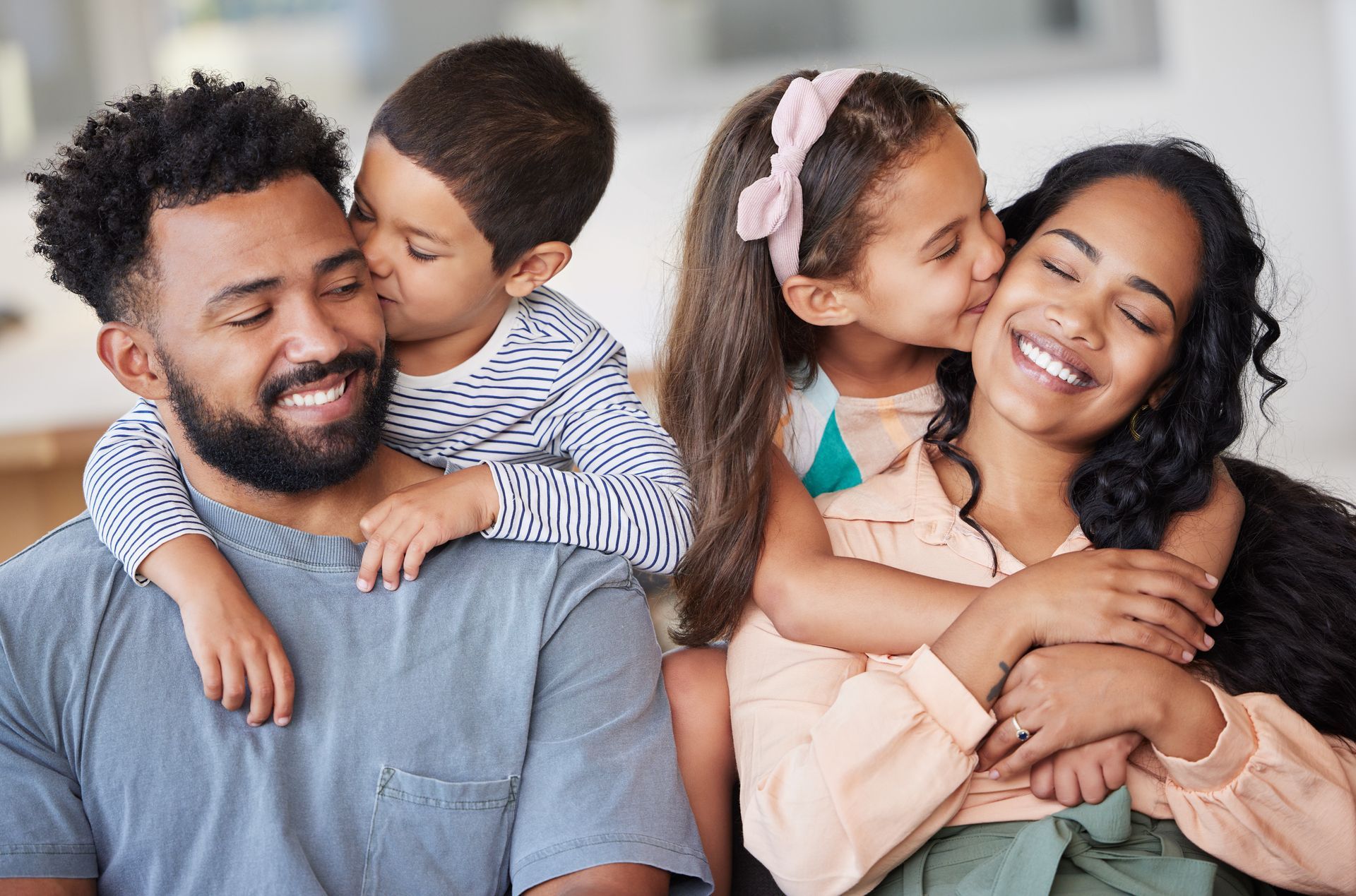 Family embraced, children kissing parents' cheeks. All smiling, light background.