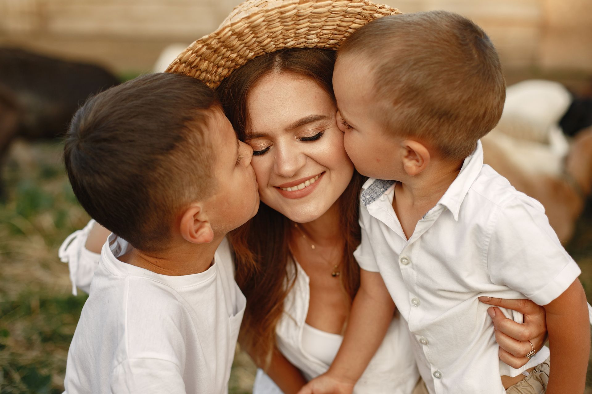 Woman wearing straw hat smiles as two children kiss her cheeks.