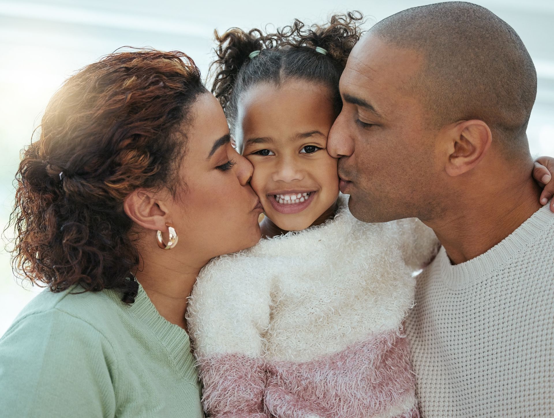 Parents kissing a child's cheeks. Family smiles for the camera, illuminated indoors.