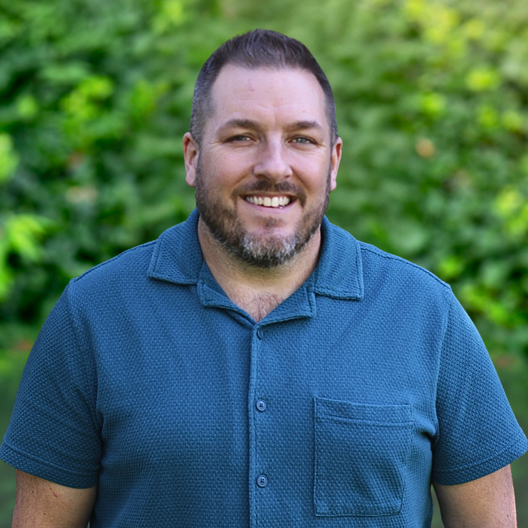 Man with dark hair and beard, wearing a blue button-down shirt, smiling.