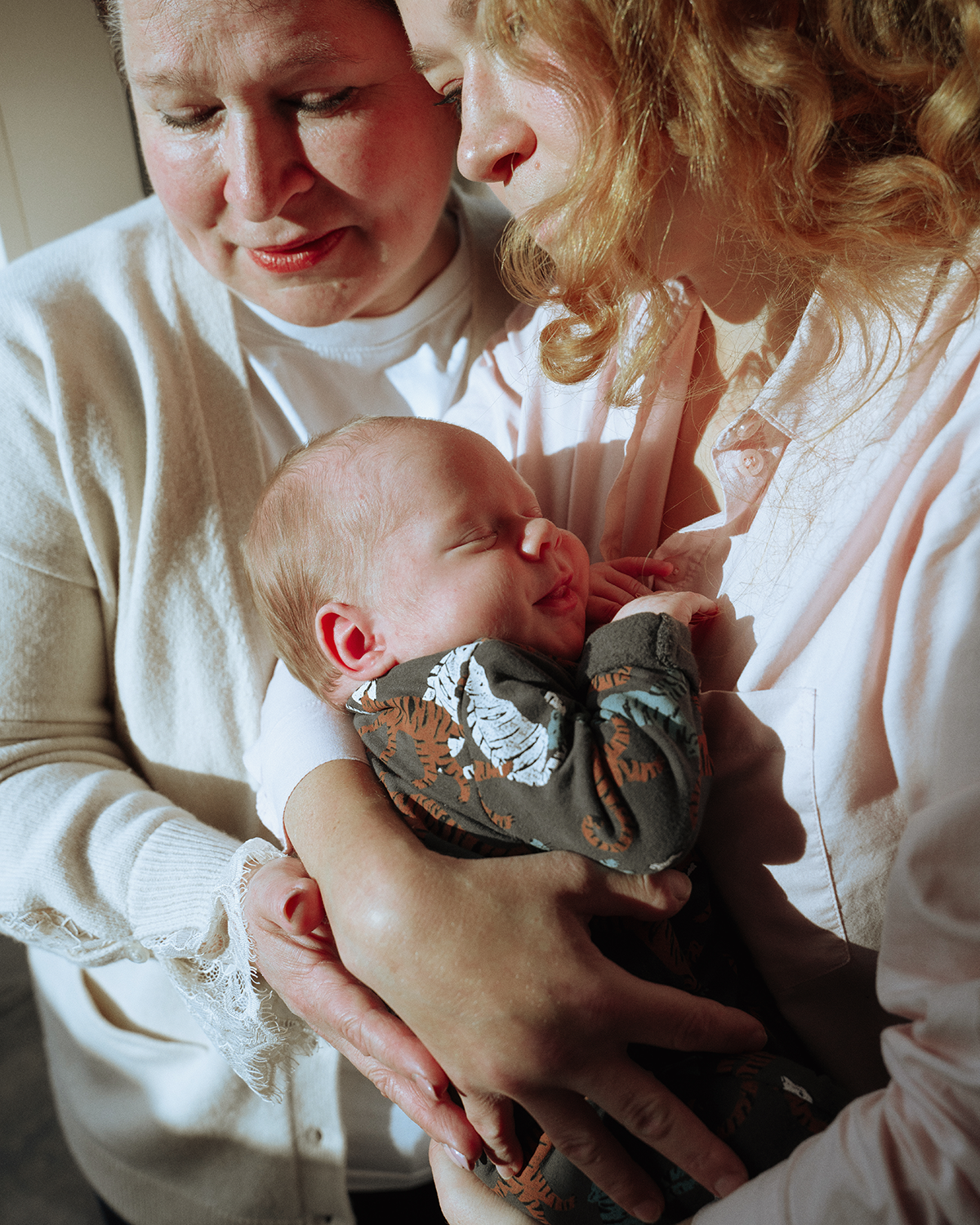 Woman holding sleeping baby, with another woman looking on. Indoors.