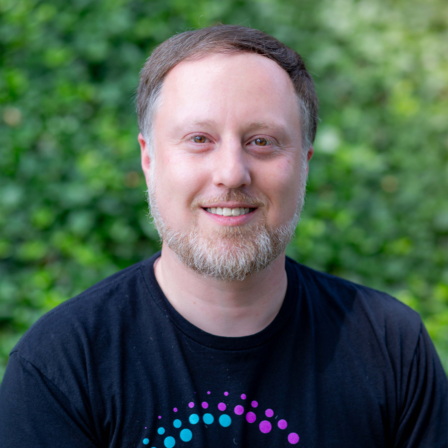 Man with light brown hair and beard, smiling, wearing a black t-shirt, in front of green foliage.