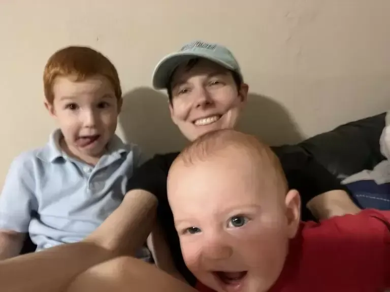 A smiling person with a light blue hat, with two children. One child is sticking out their tongue.