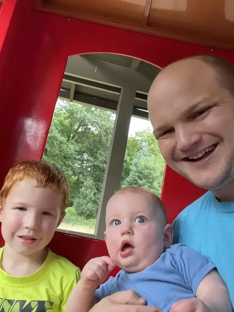 Father and two children, red-haired boy and baby, smile on a red train. Green trees visible outside the window.
