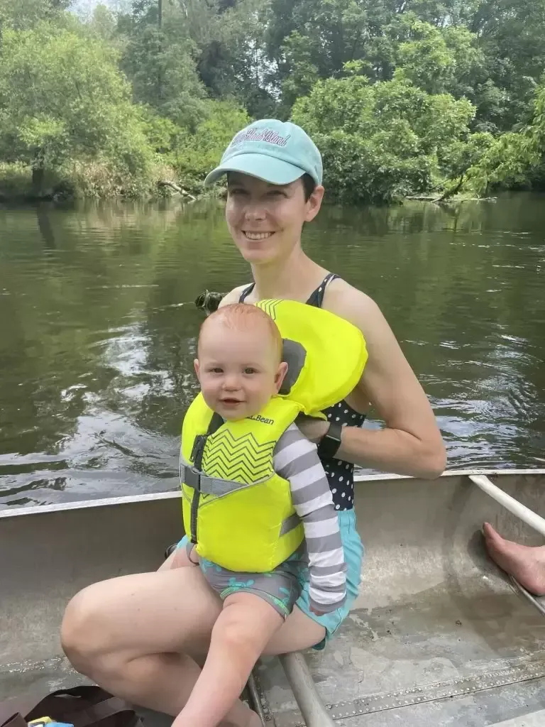 Woman in a turquoise cap and baby in a yellow life vest, smiling on a canoe trip on a river.