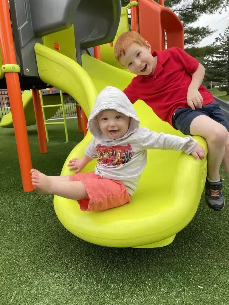 Two children on a yellow slide. One sits in the slide, smiling; the other sits above, smiling and looking. Green ground.