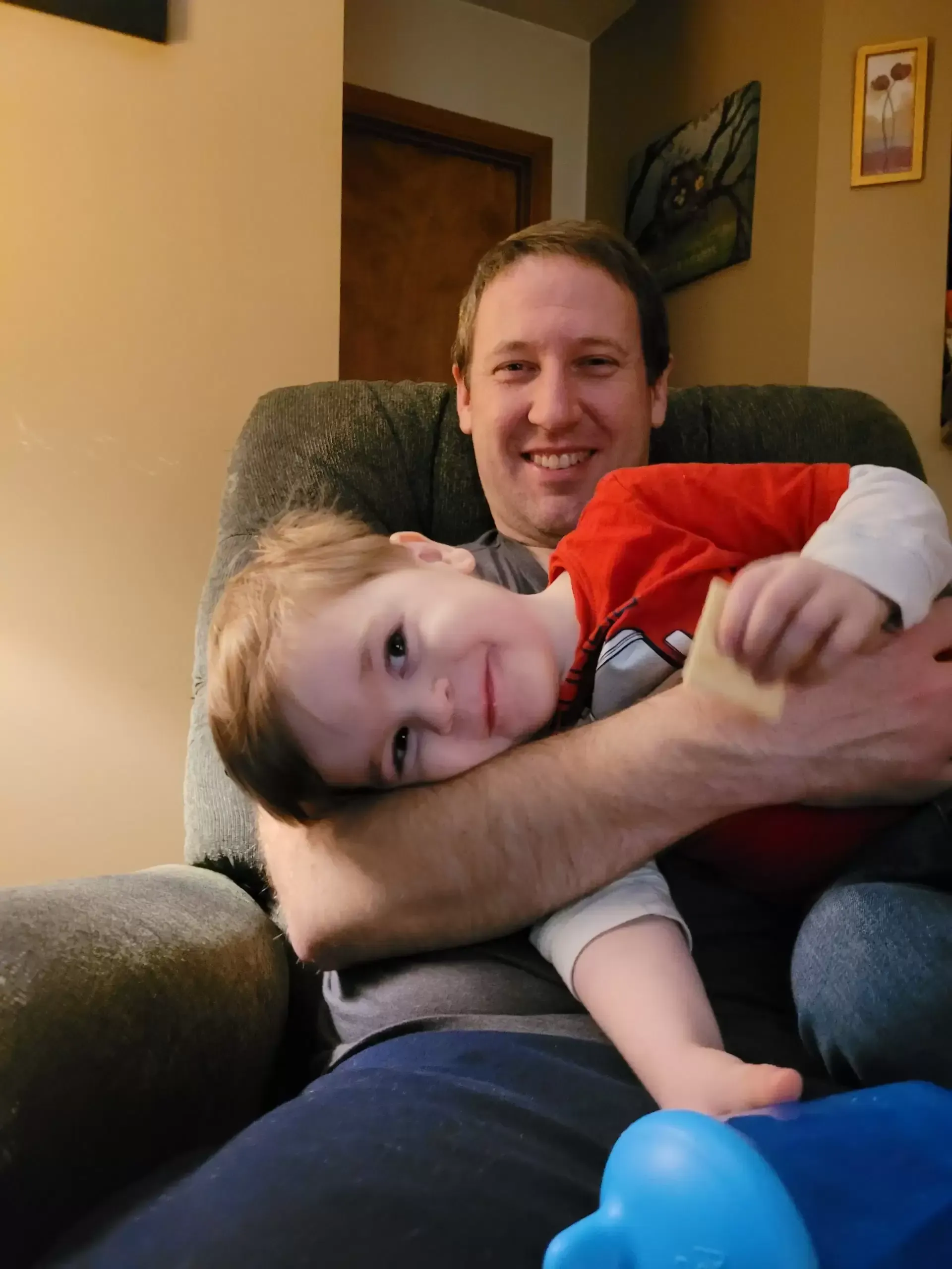 Man smiling, holding a young child. Child rests head on man's arm. Indoor setting.