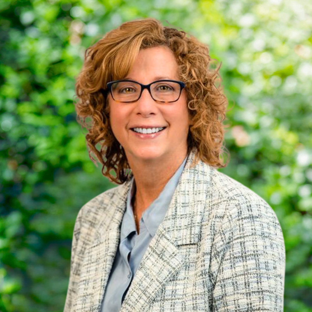 Woman with glasses smiles, wearing a dark shirt, in front of green foliage.