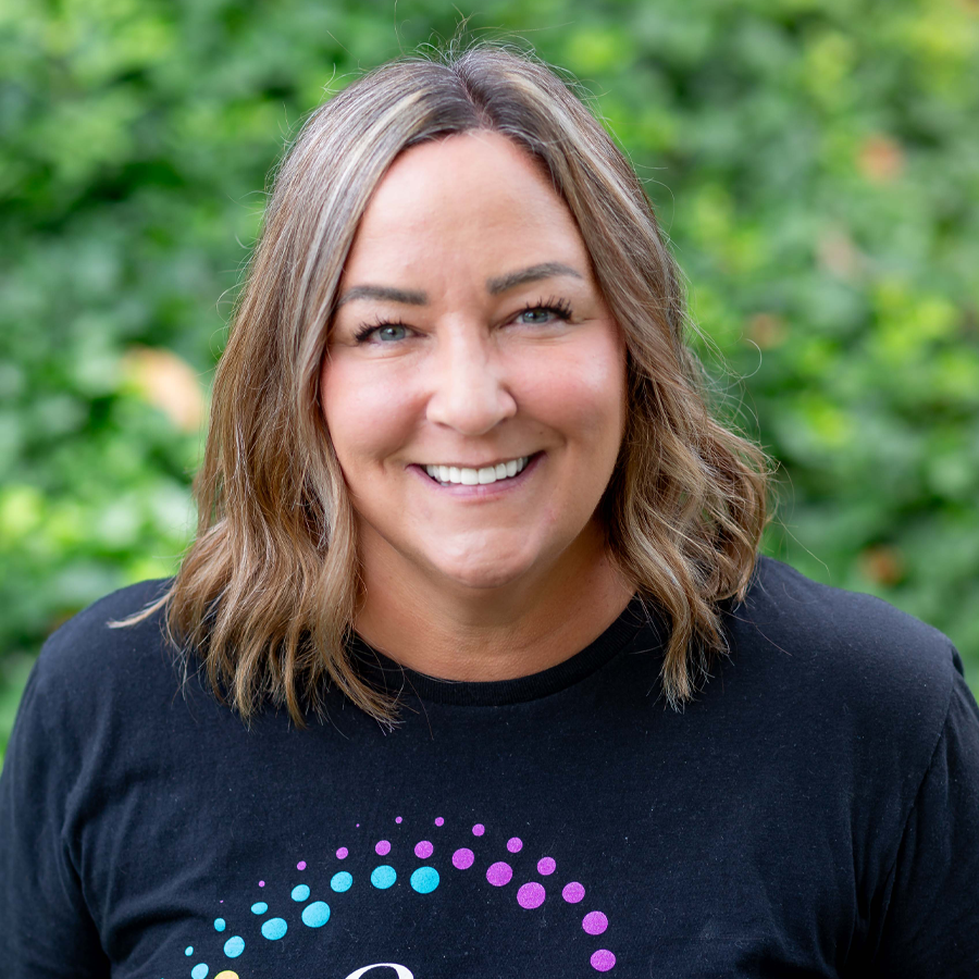 Woman with brown hair smiling outside, wearing a black shirt with a colorful design.