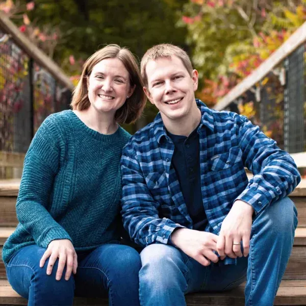 Couple sits on wooden stairs, smiling at the camera. Woman wears teal sweater, man wears blue plaid shirt. Outdoors.
