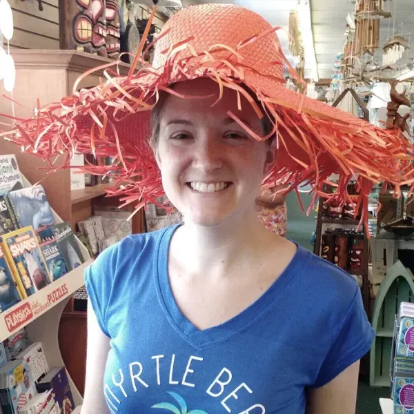 Woman wearing a large, orange, fringed hat smiles in a gift shop, wearing a blue t-shirt that says 